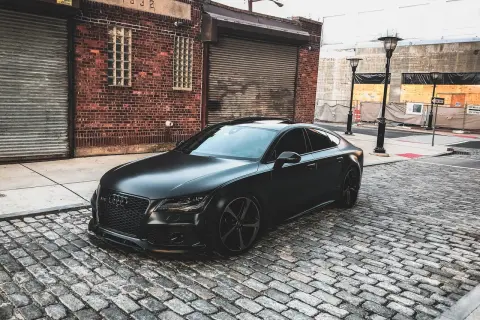 Czarny matowy samochód Audi RS7 zaparkowany na brukowanej ulicy przed ceglanym budynkiem.