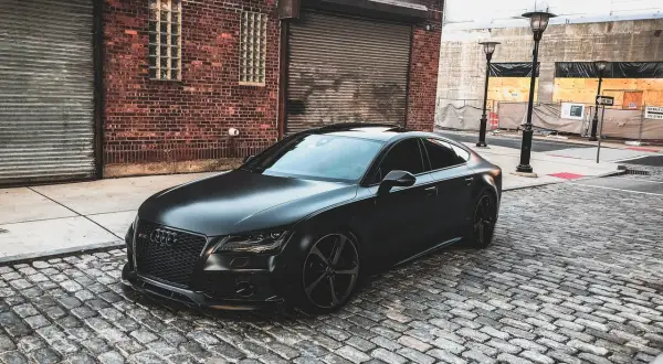 Czarny matowy samochód Audi RS7 zaparkowany na brukowanej ulicy przed ceglanym budynkiem.