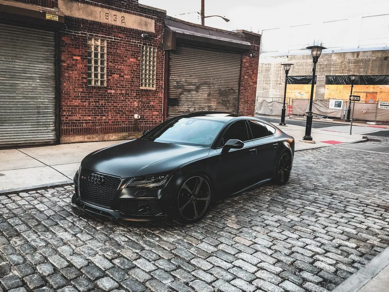 Czarny matowy samochód Audi RS7 zaparkowany na brukowanej ulicy przed ceglanym budynkiem.