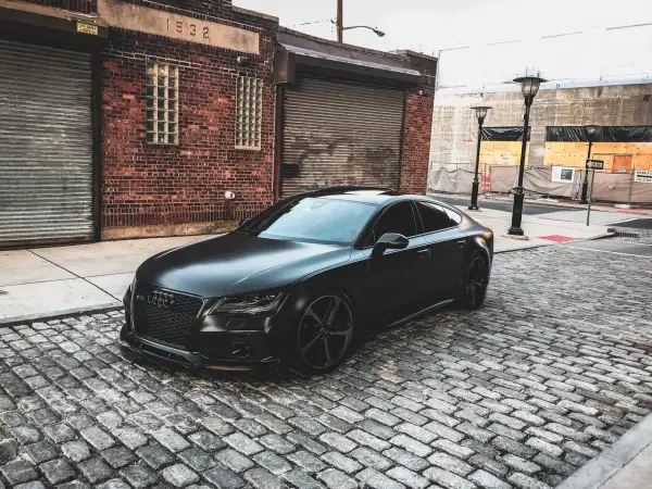 Czarny matowy samochód Audi RS7 zaparkowany na brukowanej ulicy przed ceglanym budynkiem.
